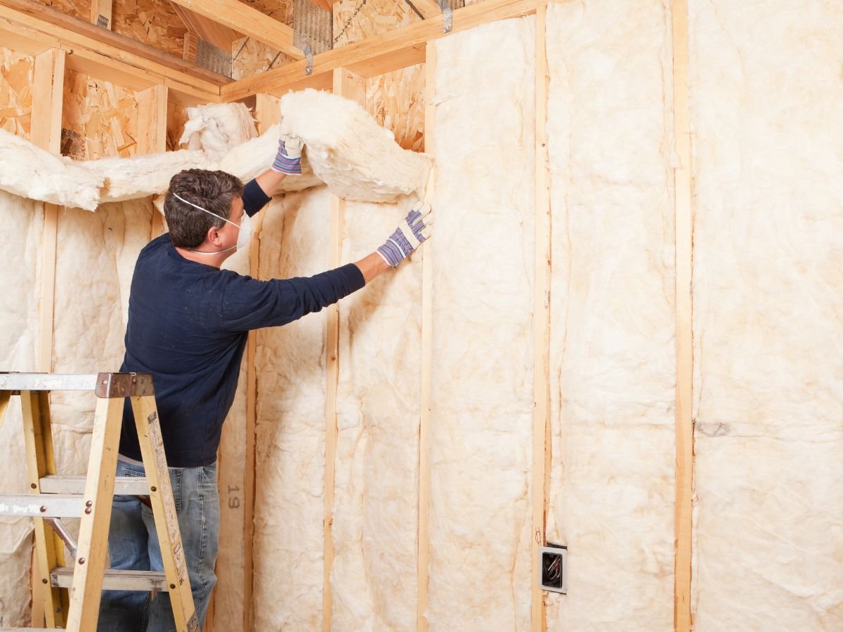 A man wearing gloves installs insulation in a wall while standing on a ladder in a wooden-framed room.